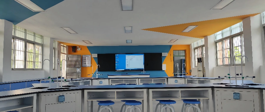 Modern school science lab with blue and white workstations, equipped with gas taps, sinks, and a fume hood, featuring a decorative wall with yellow flower patterns and a digital screen. Modern school science lab with blue and white workstations, equipped with gas taps, sinks, and a fume hood, featuring a decorative wall with yellow flower patterns and a digital screen.